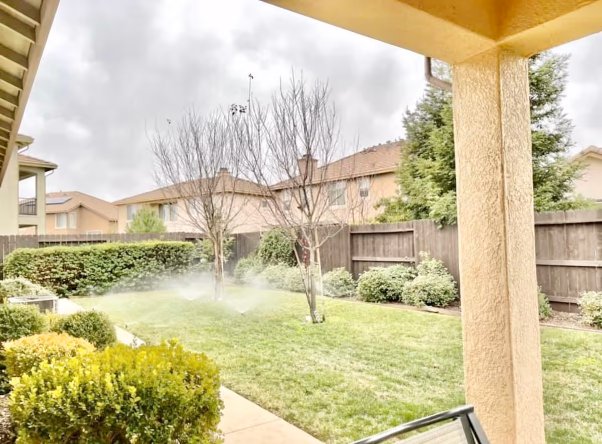 Covered patio overlooking a fenced backyard lawn with sprinklers, trees, and shrubs.