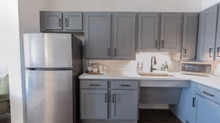 A modern kitchen with gray cabinets, a stainless steel refrigerator, a white countertop with a built-in sink, a microwave, and a white tiled backsplash.