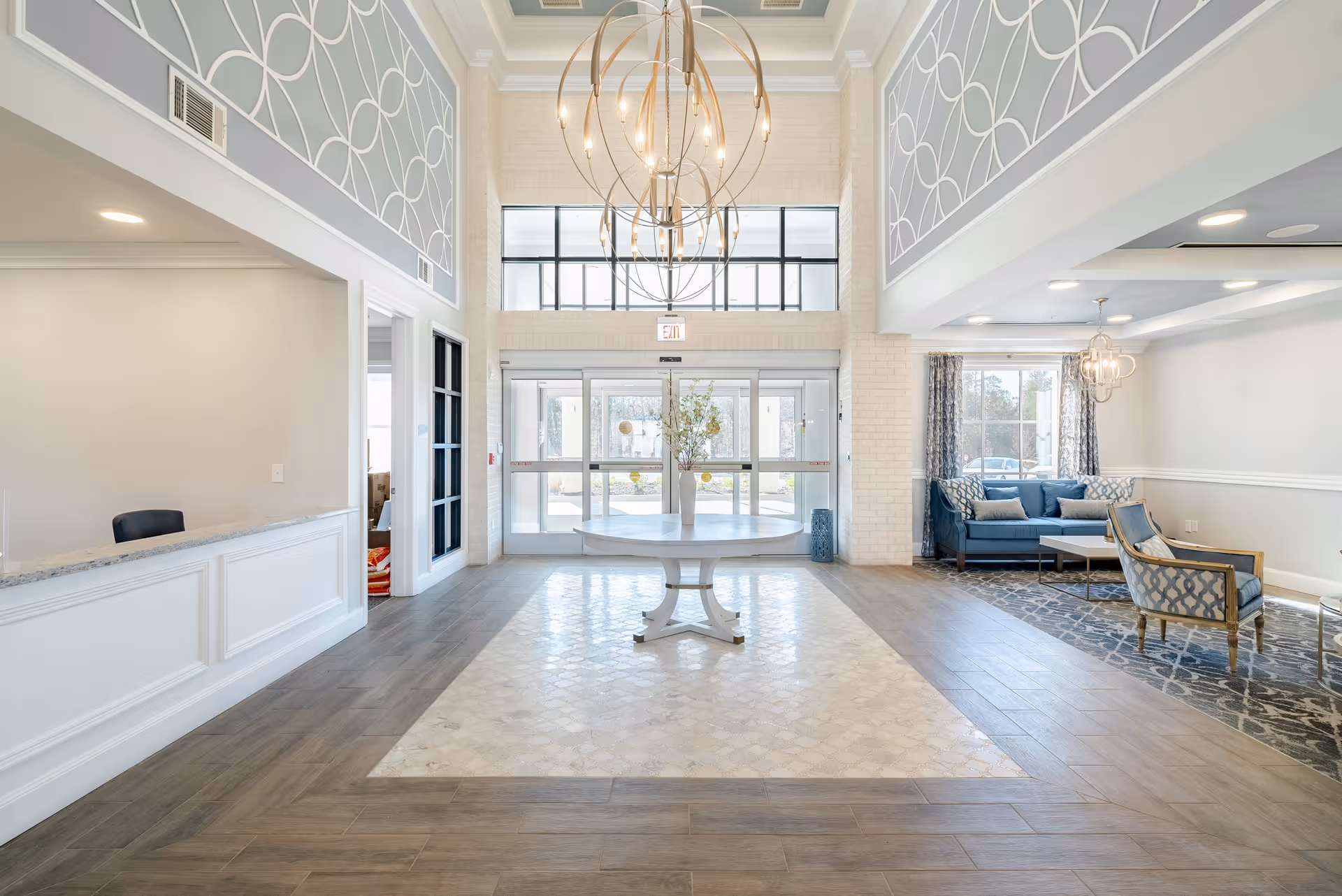 Bright, airy lobby with a central round table and chandelier, reception desk on the left and seating area on the right near the entrance doors.