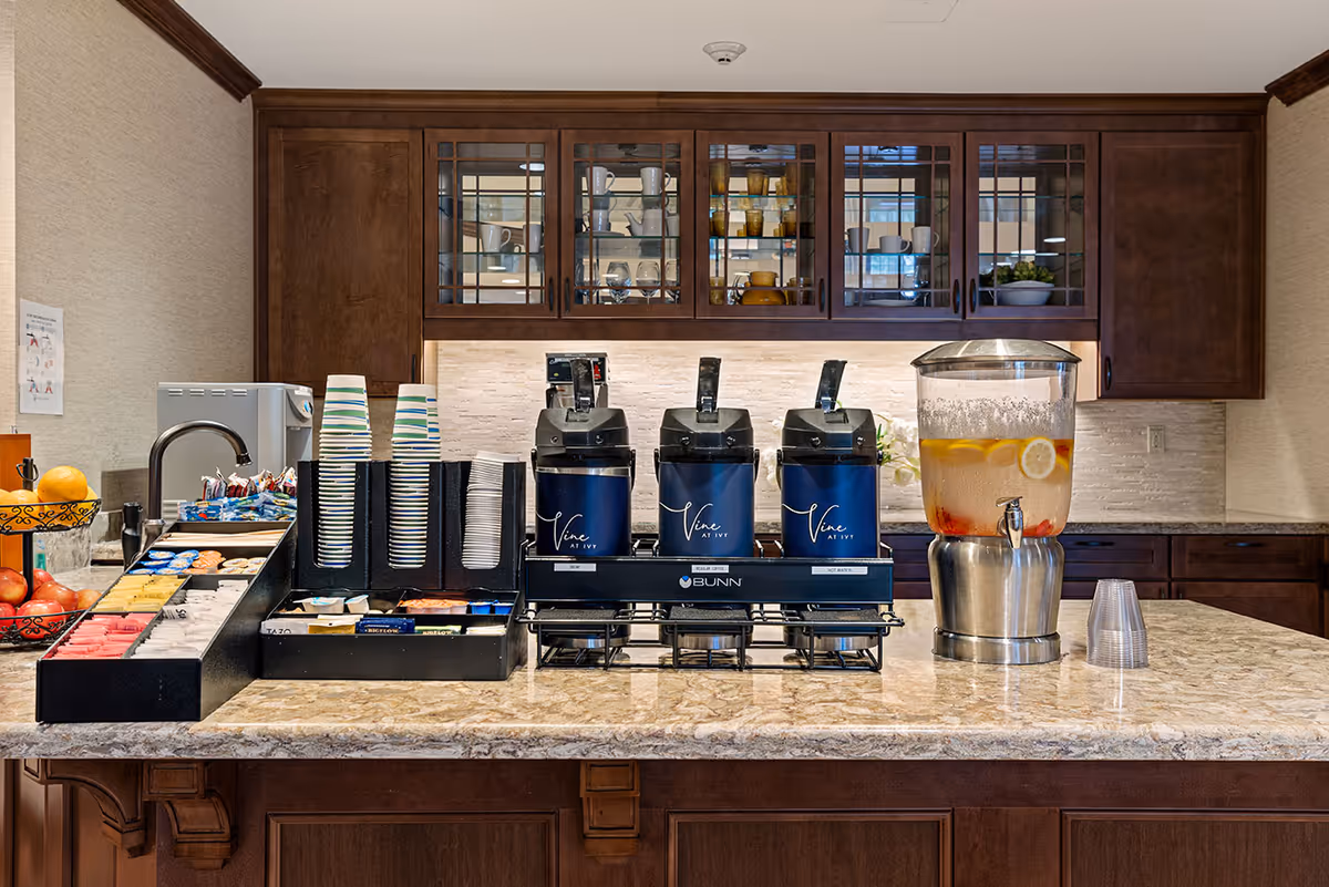 A beverage station with three coffee dispensers labeled 'Vine at Ivy,' a large glass container with infused water containing lemon and berries, stacks of disposable cups, and an assortment of tea bags and sweeteners on a marble countertop. Behind the station are wooden cabinets with glass doors displaying cups and glasses.