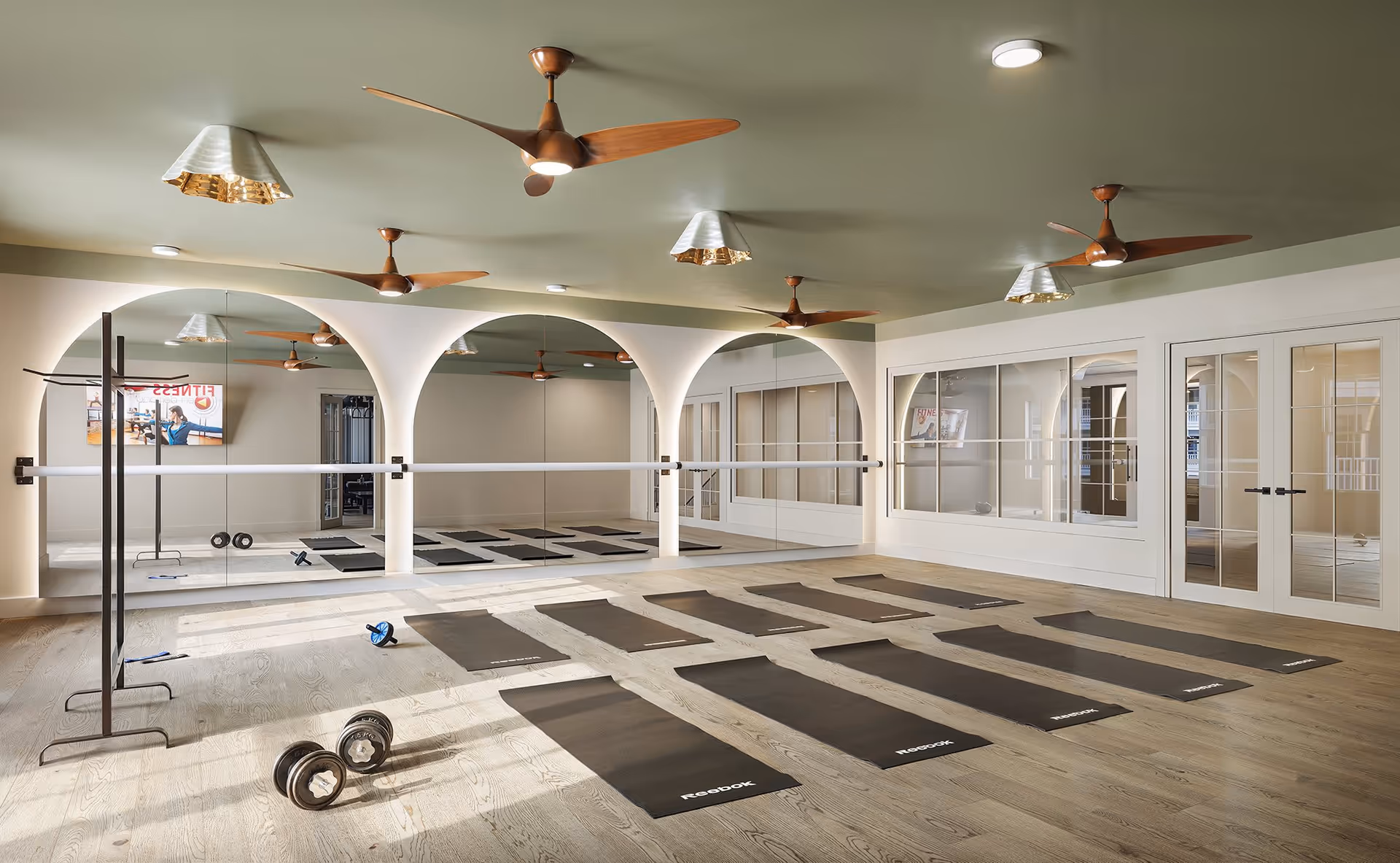 Bright fitness/yoga studio with rows of black mats, mirrors, a ballet barre, free weights and ceiling fans.