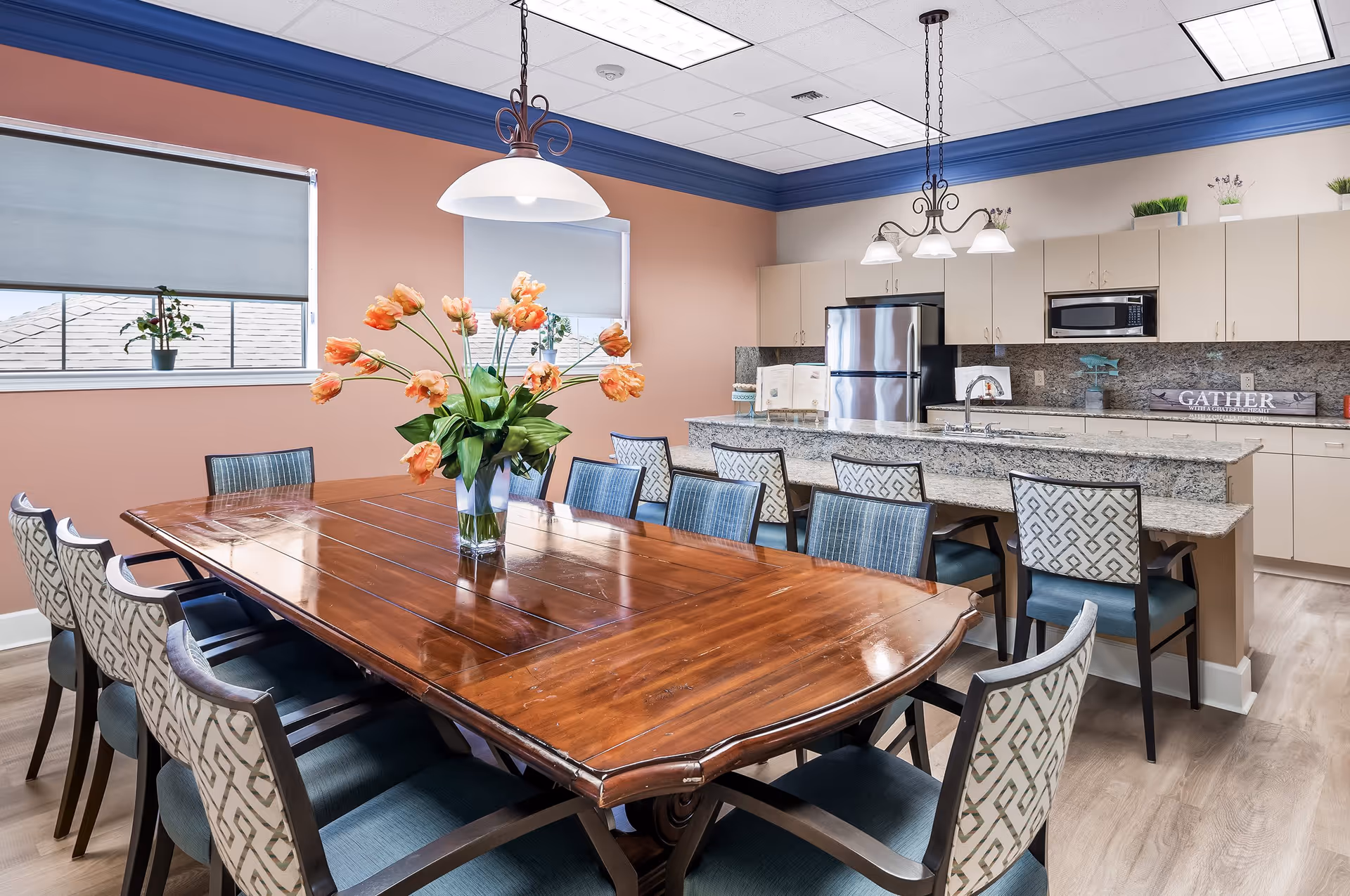 A bright and spacious dining area with a large wooden table surrounded by ten upholstered chairs. A vase with orange flowers is placed in the center of the table. In the background, there is a modern kitchen with granite countertops, stainless steel refrigerator, microwave, and beige cabinets. Two pendant lights hang above the kitchen island, and two windows with roller shades let in natural light.