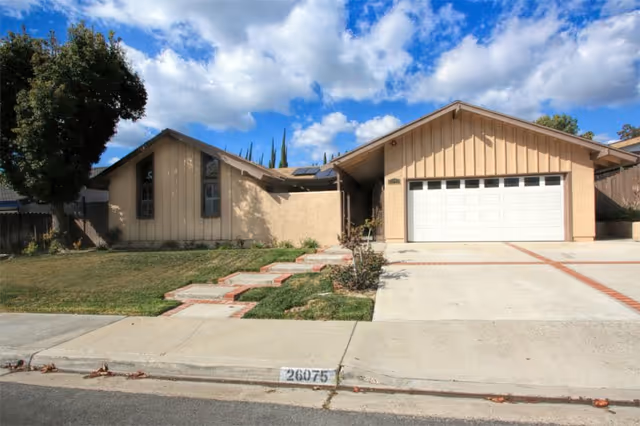 Single-story residential building with beige siding and a two-car garage under a partly cloudy blue sky. There is a concrete driveway and a stepped walkway leading to the front entrance, with a tree and some grass on the left side of the property.