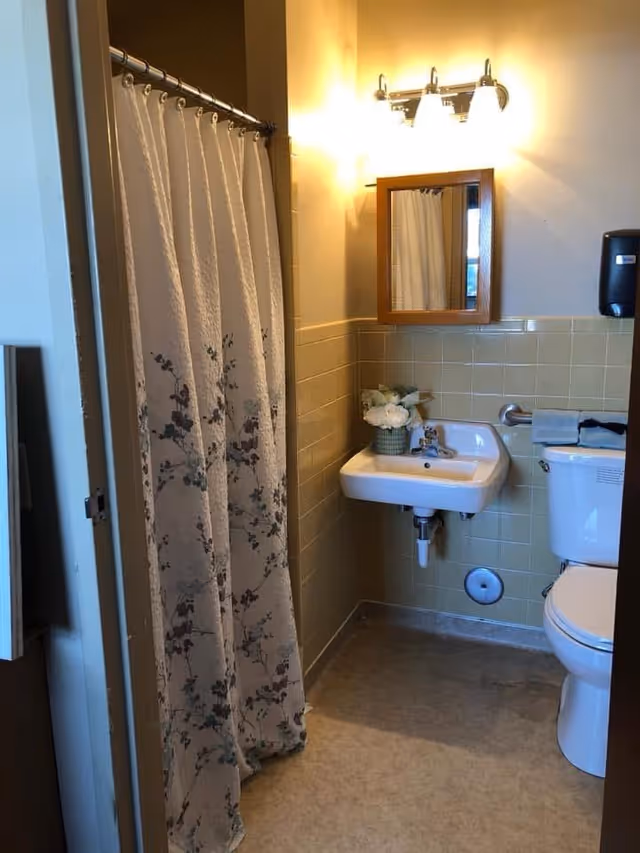 Small bathroom with a floral shower curtain, wall-mounted sink and mirror, and a toilet against yellow-green tiled walls.