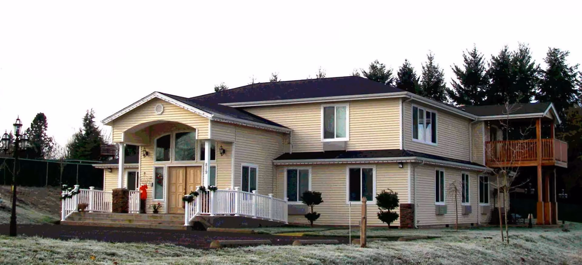 Two-story beige building with a white-railed front porch and second-floor balcony set on a frosty lawn with trees behind it.