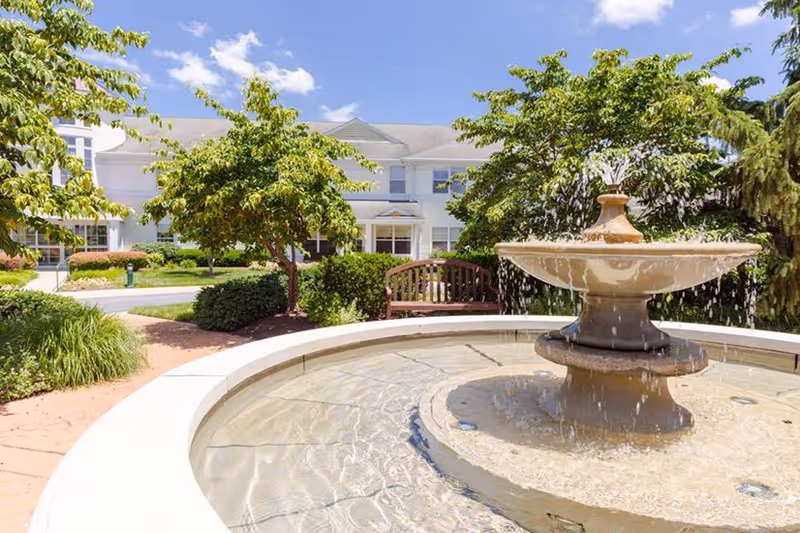 Outdoor view of Buckingham's Choice facility featuring a large circular water fountain in the foreground, surrounded by greenery including trees and bushes, with a wooden bench and the building visible in the background under a blue sky with some clouds.