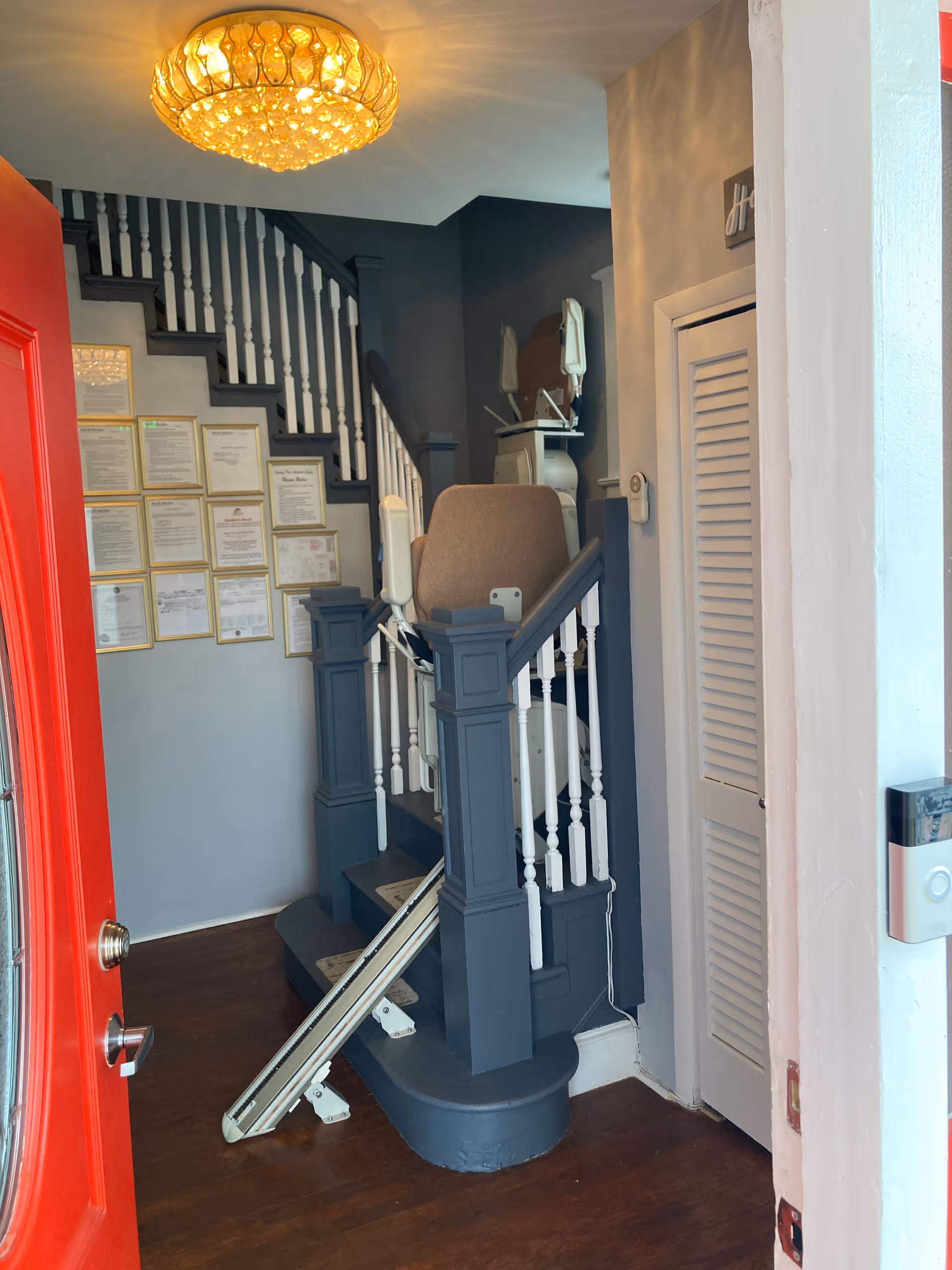 View of an interior stairway with a stair lift installed on the right side. The stairs have dark blue and white railings, and there are multiple framed certificates or documents on the wall beside the stairs. A bright chandelier hangs from the ceiling, and a red door is partially open in the foreground.