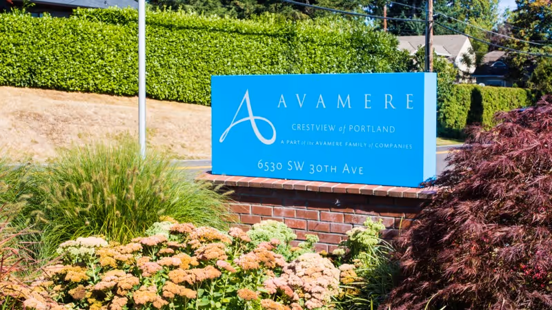 Blue sign for Avamere Crestview of Portland located outdoors surrounded by green bushes, ornamental grasses, and flowering plants, with a brick base and residential houses in the background.