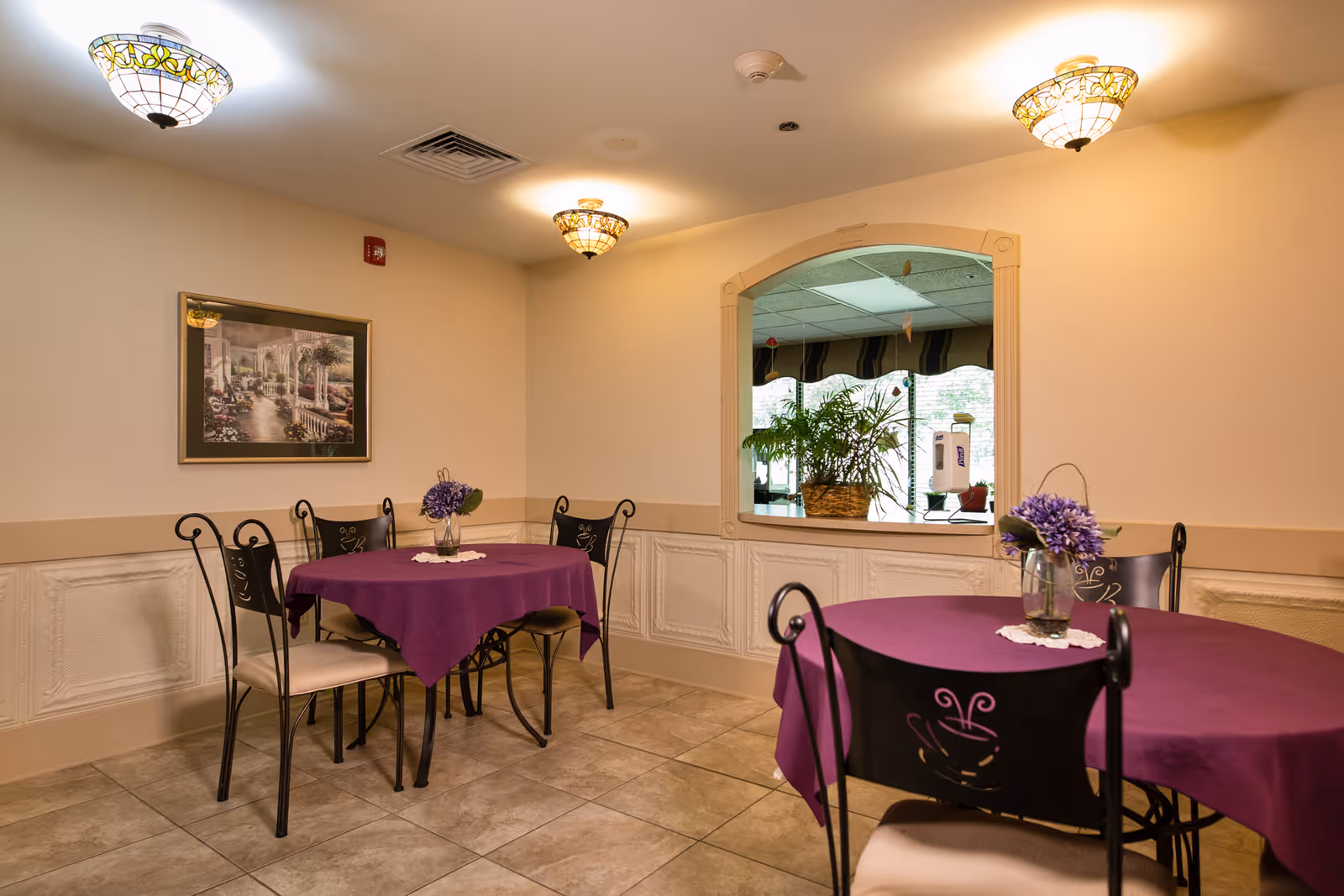 Dining area with round tables draped in purple tablecloths, wrought-iron chairs, a framed painting, and a passthrough window with plants.