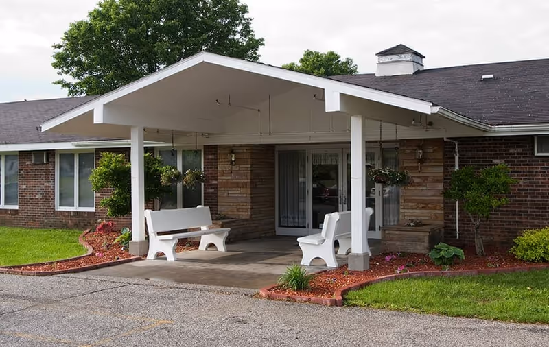 Entrance of a nursing and rehabilitation facility with a covered porch supported by white pillars, two white benches, hanging flower baskets, and landscaped greenery with grass and small bushes.
