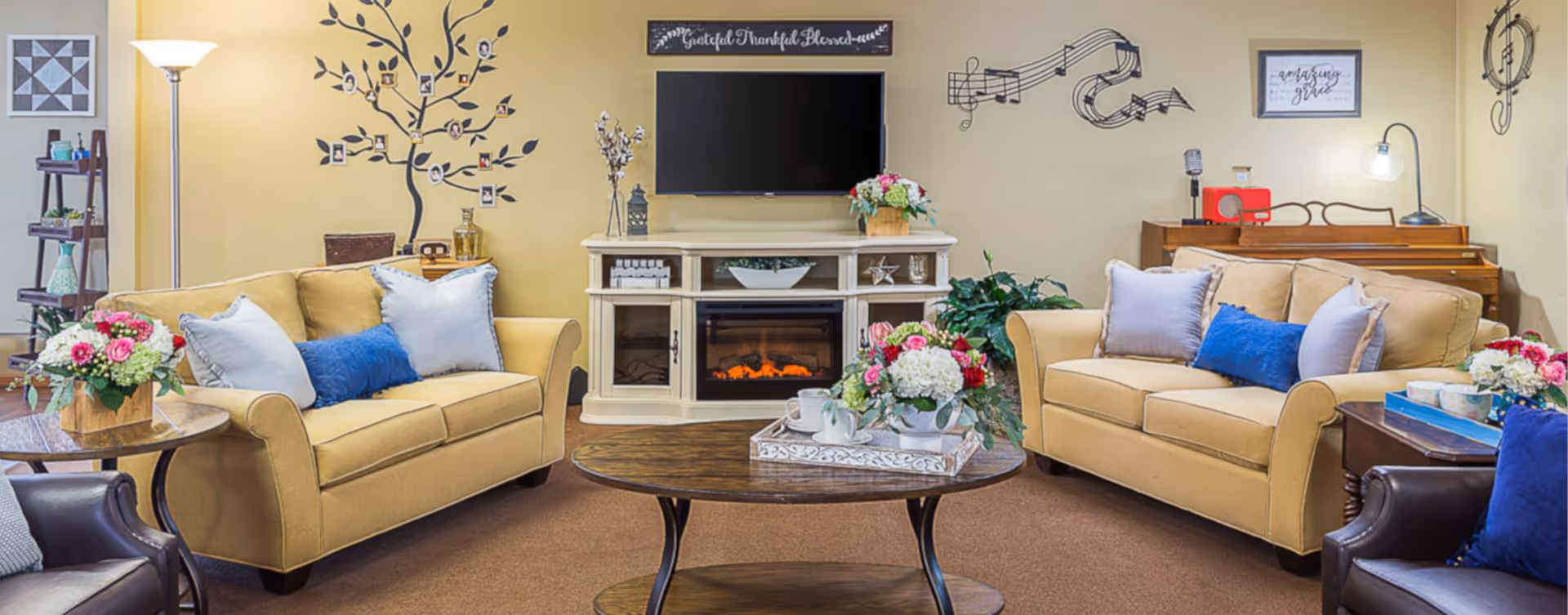 A cozy living room with two beige sofas adorned with blue and white pillows, a wooden coffee table with floral arrangements and tea set, a white electric fireplace with a TV mounted above it, and decorative wall art including a family tree and musical notes. There is a floor lamp, a wooden piano with a lamp and a red radio, and additional seating with a dark leather chair.