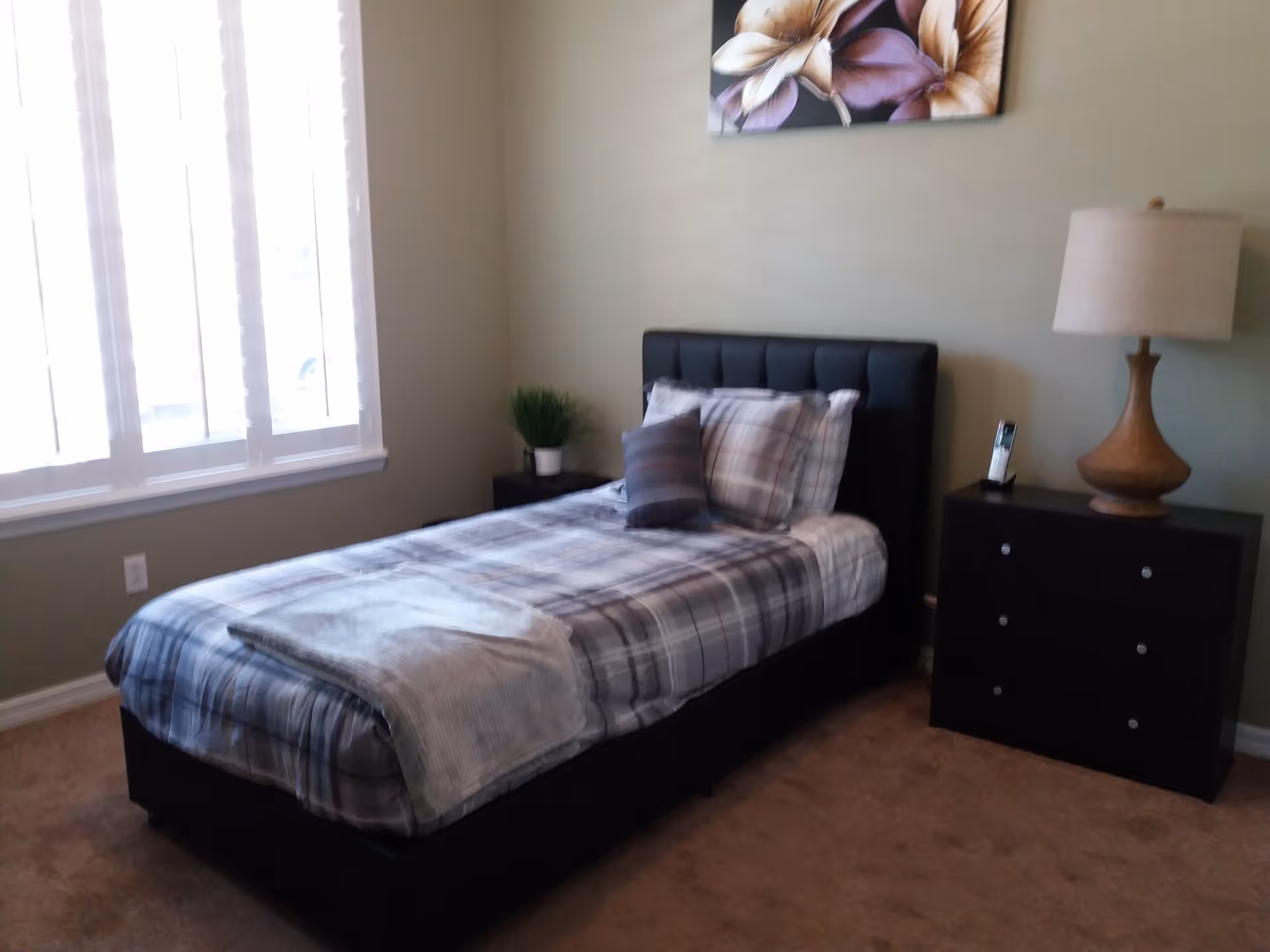 A bedroom with a single bed featuring a plaid comforter and pillows. Next to the bed is a dark wooden nightstand with a lamp and a phone. A small plant is on a side table near the window with white blinds. A floral painting hangs on the wall above the bed.