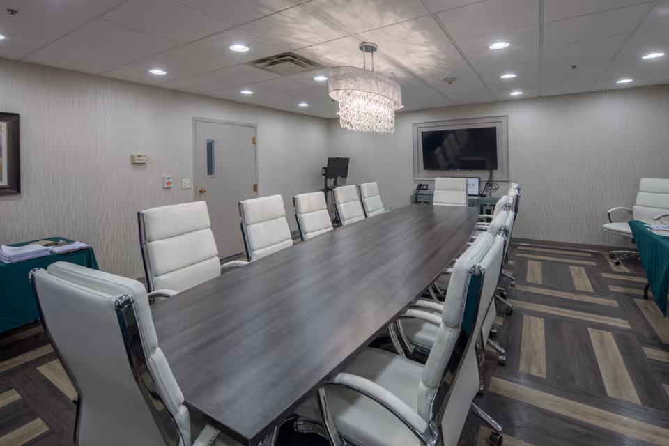 Conference/meeting room with a long dark table surrounded by white leather chairs, a chandelier, and a wall-mounted TV.