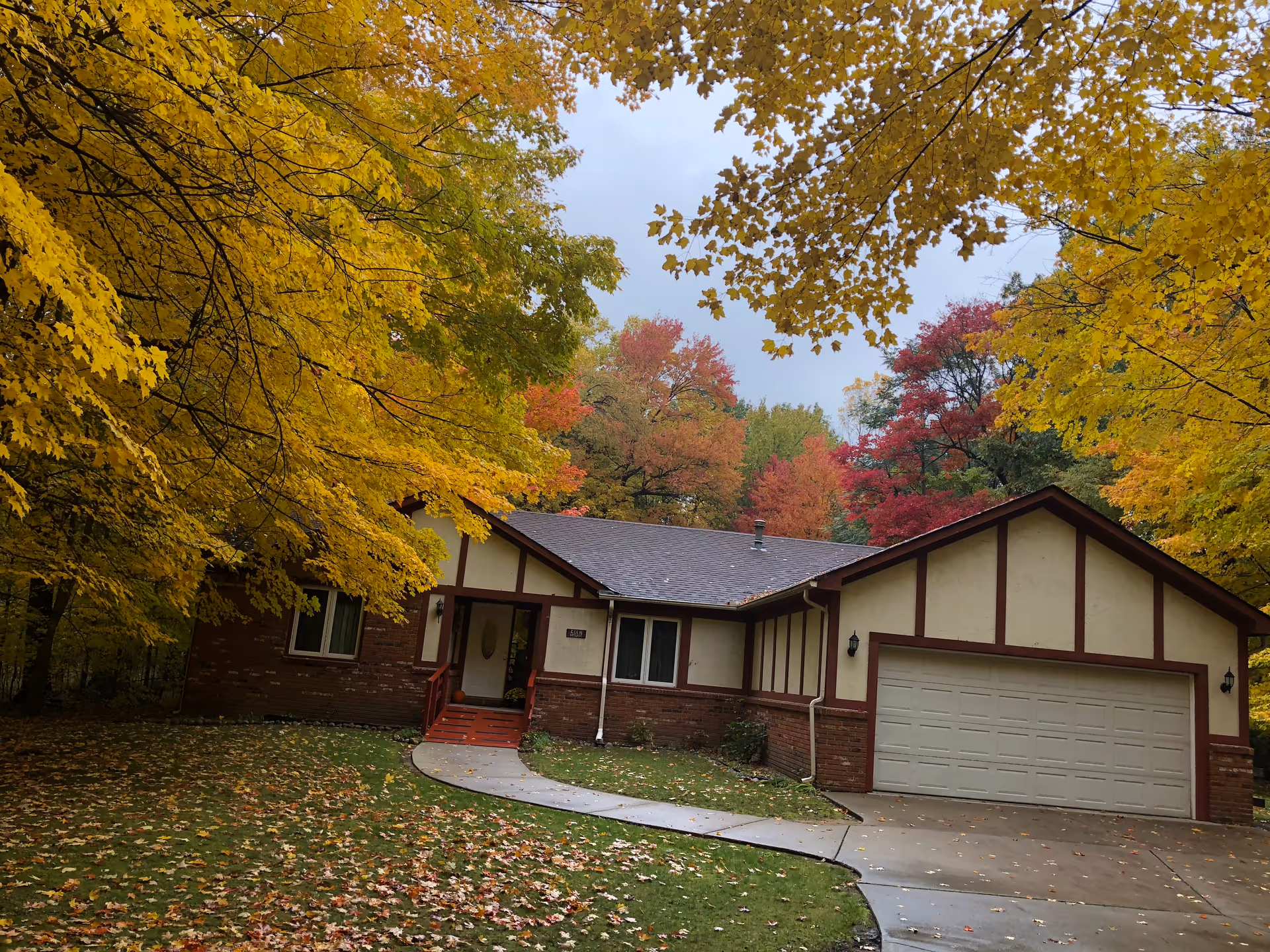 A single-story house with a two-car garage surrounded by trees with autumn foliage in shades of yellow, red, and green. The house has a brick and beige exterior with brown trim, a curved concrete walkway leading to the front door, and a lawn covered with fallen leaves.