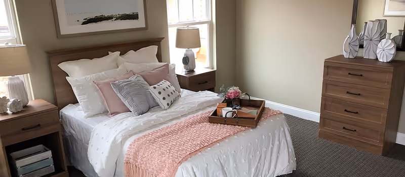 A neatly made bedroom with a wooden bed, bedside tables and lamps, a dresser, and decorative pillows and a pink throw.