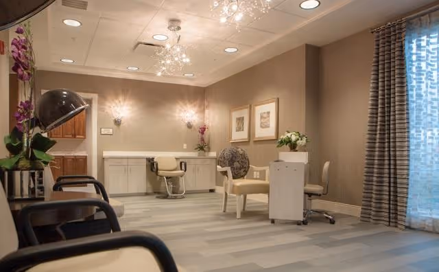 A well-lit interior room with beige walls and light gray flooring, featuring salon-style chairs, a hair dryer hood, a vanity with a chair, and decorative plants and framed pictures on the walls. A window with patterned curtains allows natural light to enter the space.