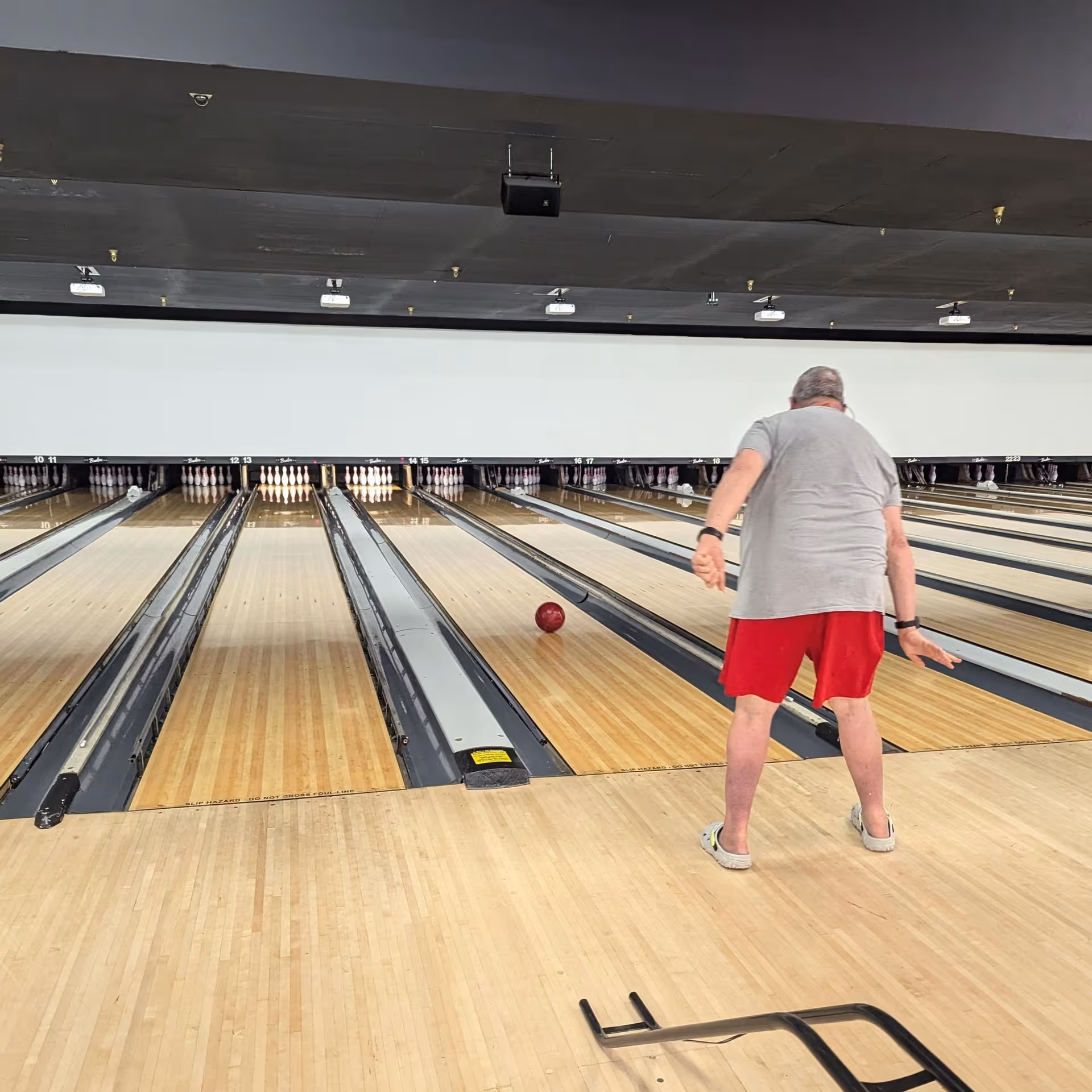A person in a gray shirt and red shorts rolls a bowling ball down an indoor bowling lane toward the pins.