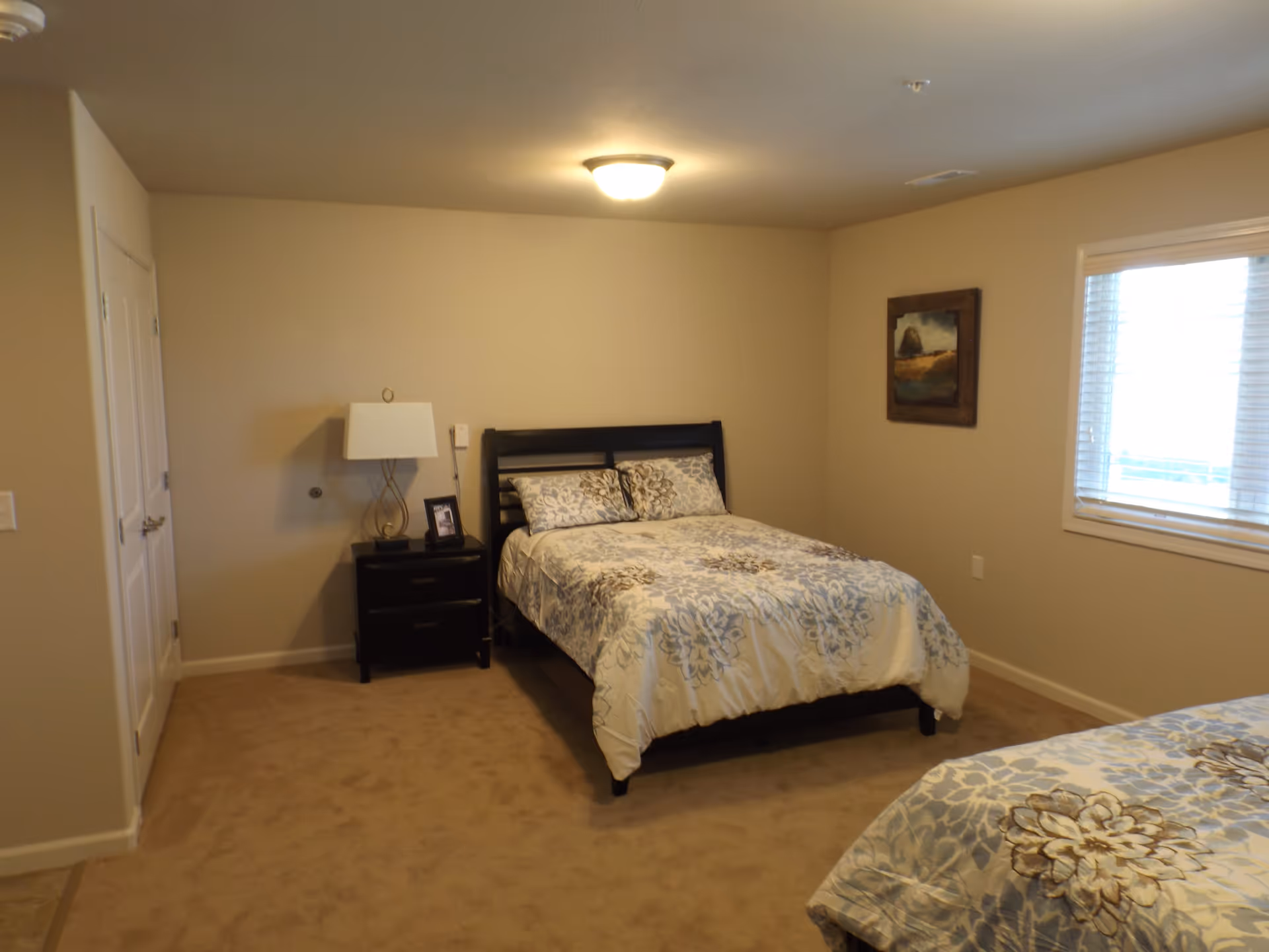 A bedroom with beige walls and carpet, featuring a bed with a floral patterned comforter in shades of blue and beige. There is a nightstand with a lamp and a framed photo next to the bed. A window with white blinds is on the right wall, and a framed painting hangs between the bed and the window. A closet door is visible on the left side of the room.