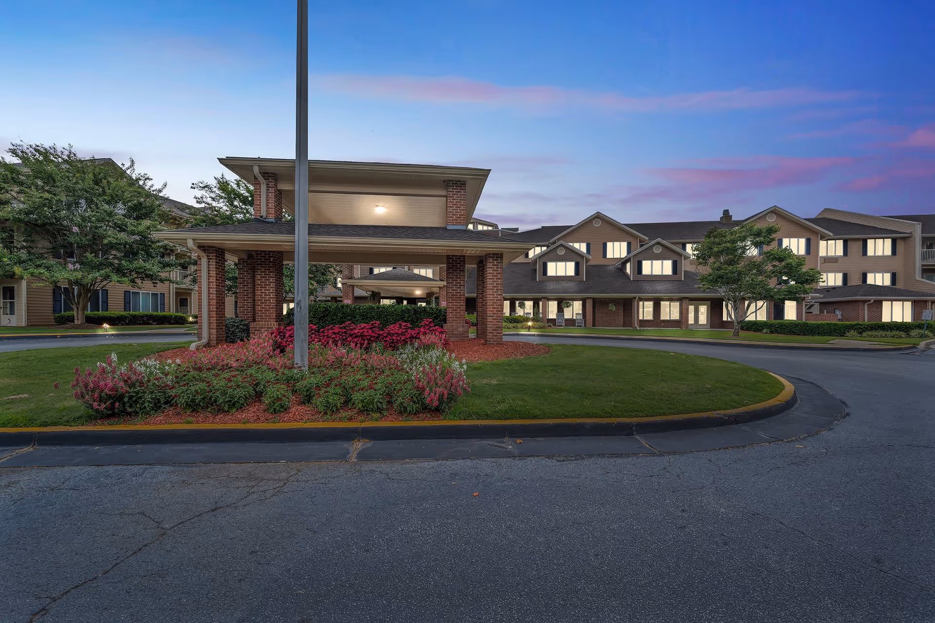 Exterior view of Holiday Pinegate senior living facility at dusk, showing a covered entrance with brick pillars, landscaped flower beds, green lawn, and a multi-story building with lit windows in the background under a blue and purple sky.