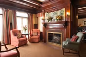 A cozy living room with wooden paneling and a fireplace. The room features two pink upholstered armchairs with a small round table between them, a green armchair with a red pillow, floral curtains, and decorative items on the mantel above the fireplace.