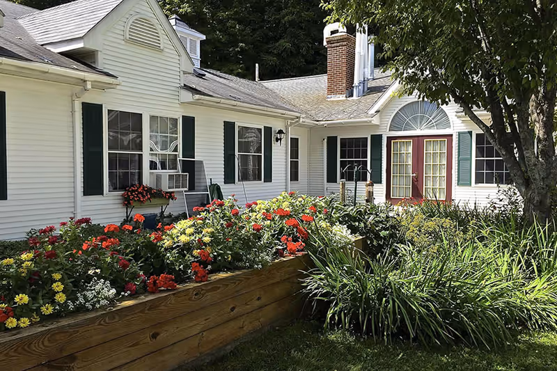 Exterior view of a white building with green shutters and a red door, surrounded by a garden with colorful flowers and greenery under a sunny sky.