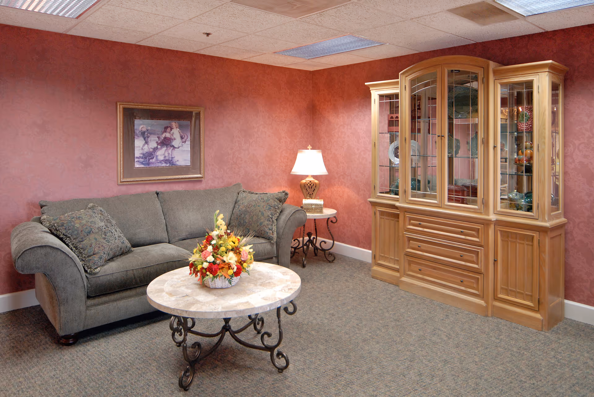 A cozy living room with a green upholstered sofa, round marble-top coffee table topped with a floral arrangement, a side table with a lamp, and a wooden display cabinet against a red patterned wall.
