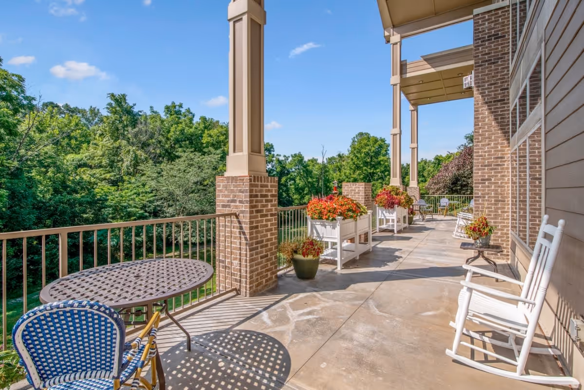 A spacious outdoor patio area with a round metal table and blue woven chair, white rocking chairs, and several planters filled with red and orange flowers. The patio is supported by brick columns and overlooks a lush green wooded area under a clear blue sky.