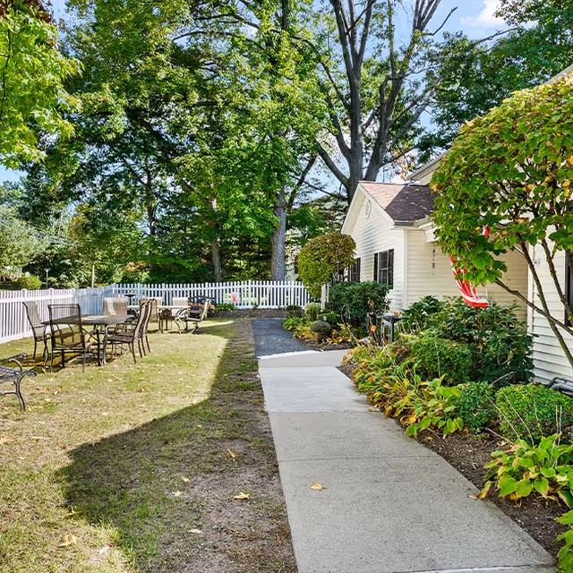 Outdoor garden area at Fox Trail Memory Care Living featuring a paved walkway alongside a white building with black shutters, surrounded by green shrubs and trees. There are several metal tables and chairs on the grass near a white picket fence under a clear blue sky.