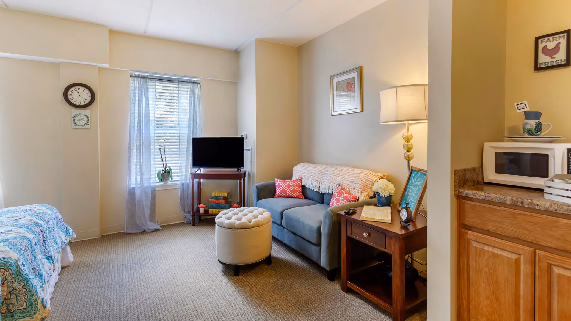A cozy living area in a senior living facility featuring a blue loveseat with red patterned pillows, a round beige ottoman, a wooden side table with a lamp, flowers, and a small clock. To the left, there is a bed with a blue and white patterned bedspread. A window with sheer curtains lets in natural light, and a small TV sits on a wooden stand with books underneath. On the right, a countertop with a microwave and decorative items is visible.
