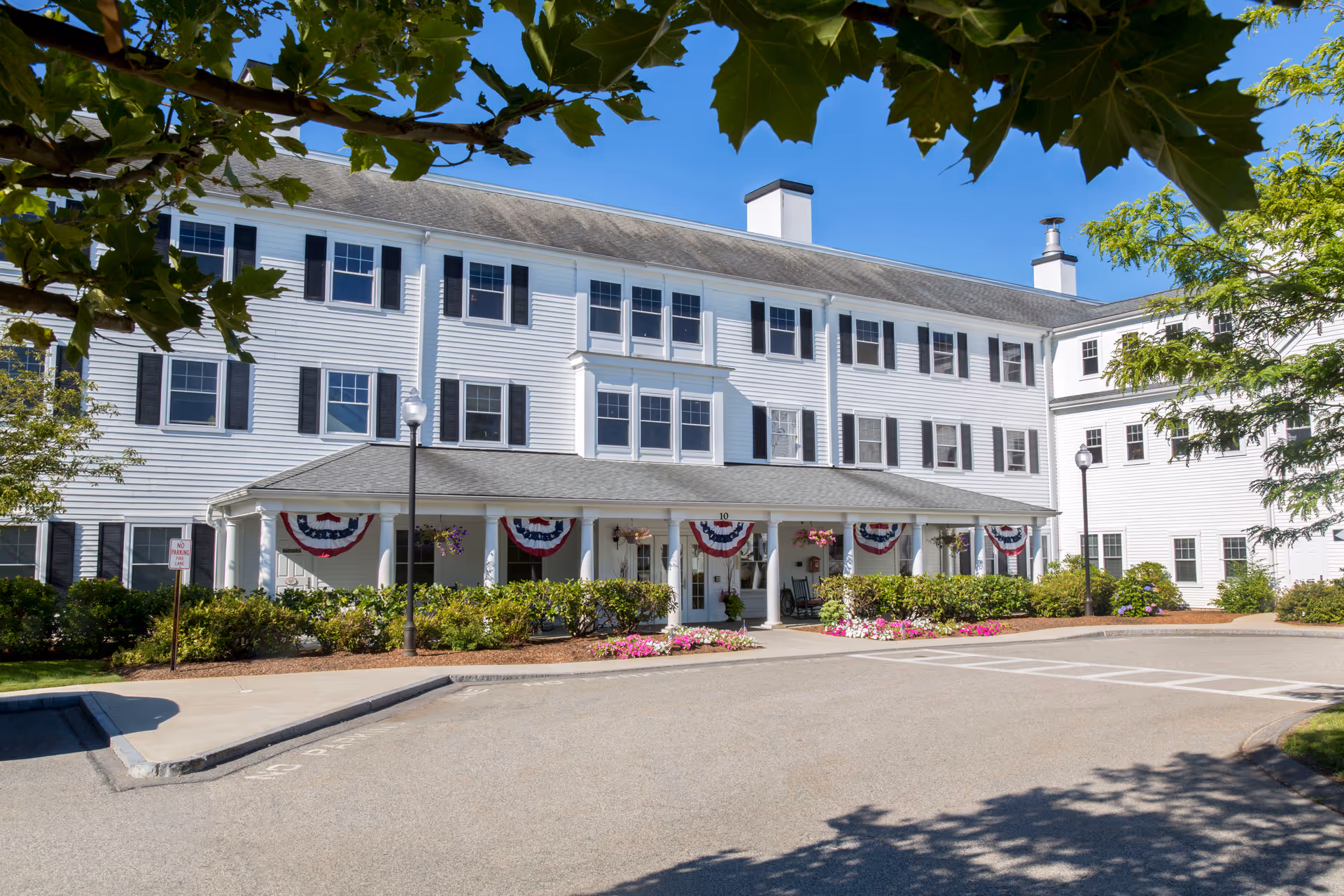 Exterior view of a three-story white senior living facility building with black shutters and a covered entrance decorated with red, white, and blue bunting. The building is surrounded by greenery and flowers, with a clear blue sky overhead.
