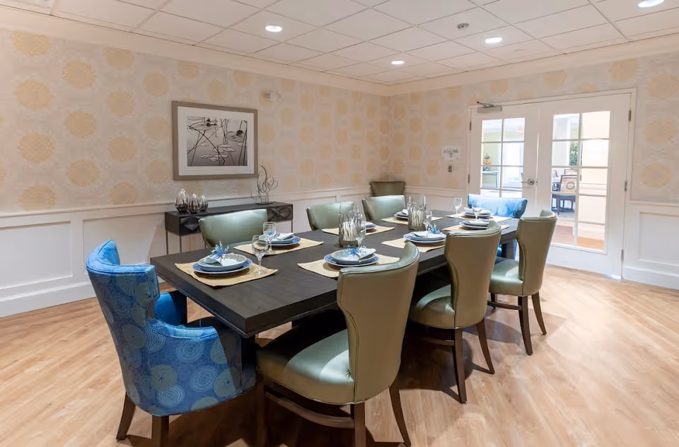 A dining room with a dark wooden rectangular table set for eight people. The table has placemats, plates, napkins with decorative holders, and wine glasses. There are six green upholstered chairs and two blue patterned armchairs around the table. The room has light wood flooring, patterned wallpaper, white wainscoting, and a framed black and white artwork on the wall. Double glass doors lead to another room.