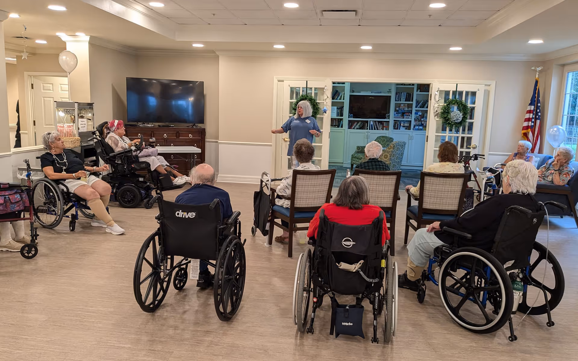 A group of elderly individuals, some in wheelchairs, seated in a semi-circle facing a woman standing and speaking in a common area of a senior living facility. The room has light-colored walls, a large TV mounted on a dresser, and a bookshelf with decorations including wreaths. An American flag is visible near a window on the right side.