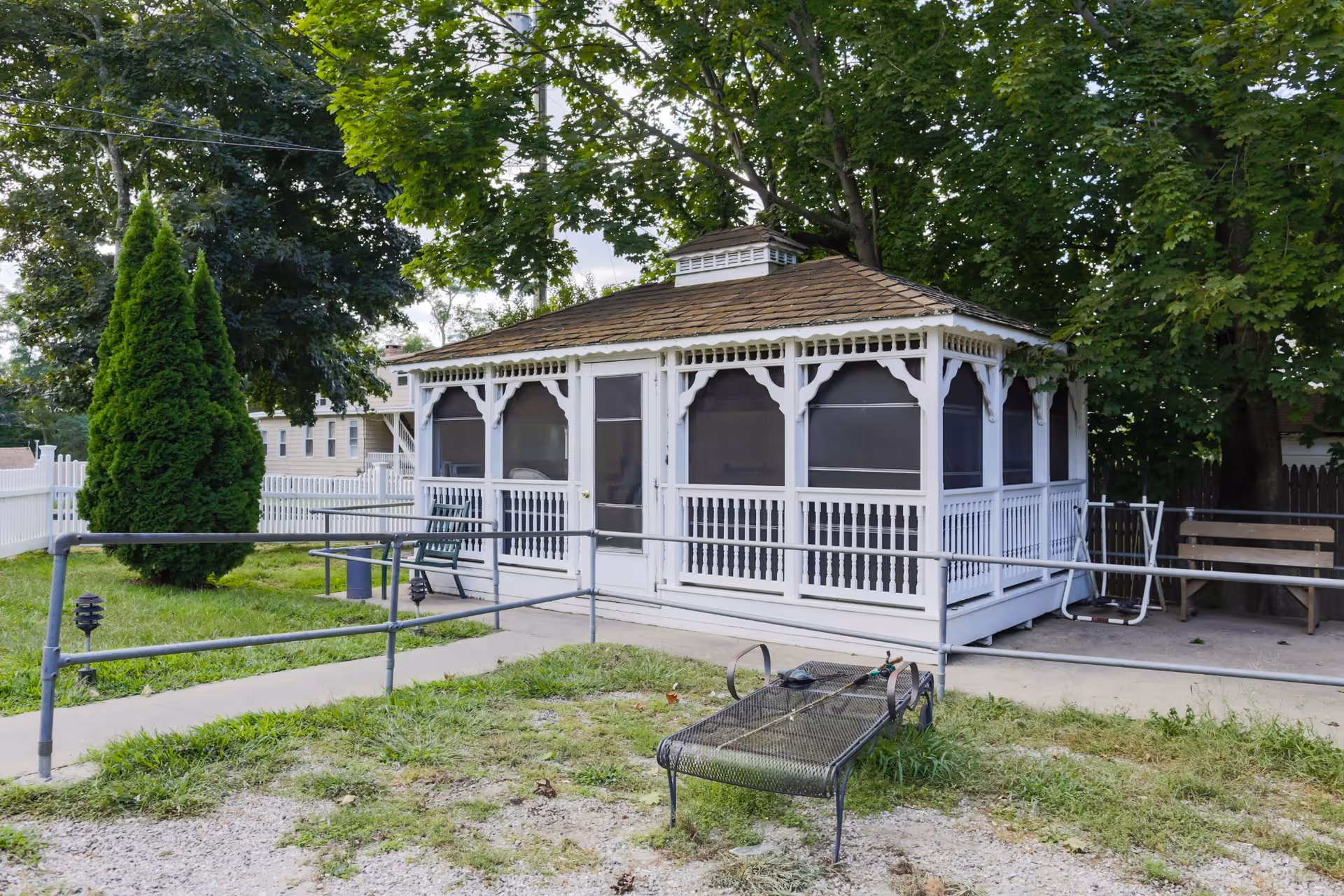 A white gazebo with screened windows and decorative trim situated in a grassy outdoor area with trees and a white picket fence in the background. There is a metal bench and a wooden bench nearby, along with a paved walkway and metal handrails.