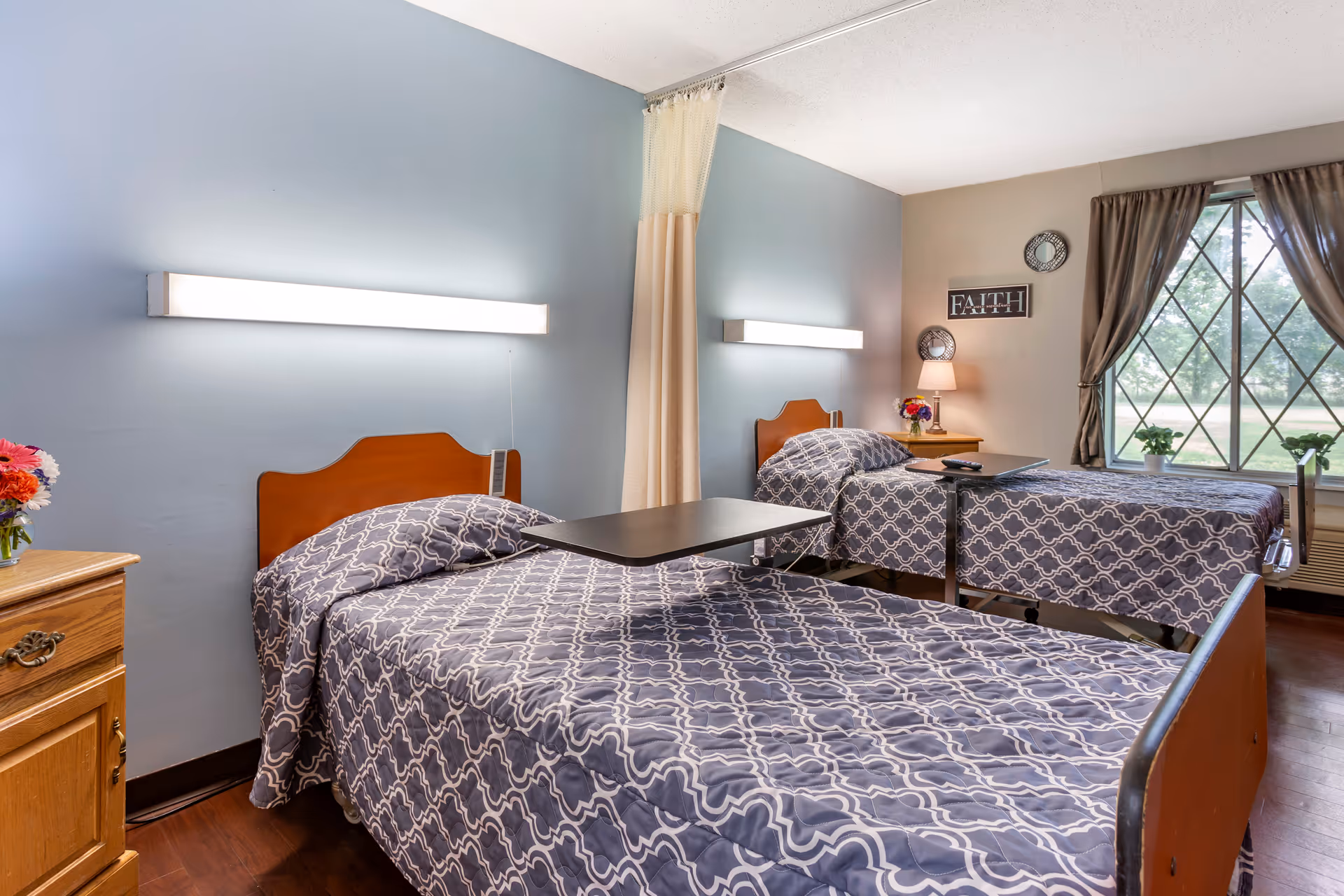 A bright and clean room with two single beds, each covered with matching gray and white patterned bedspreads. Each bed has a wooden headboard and an adjustable overbed table. The room has light blue and beige walls, a window with brown curtains, a small wooden nightstand with a lamp and flowers, and a wall decoration that says 'FAITH'.