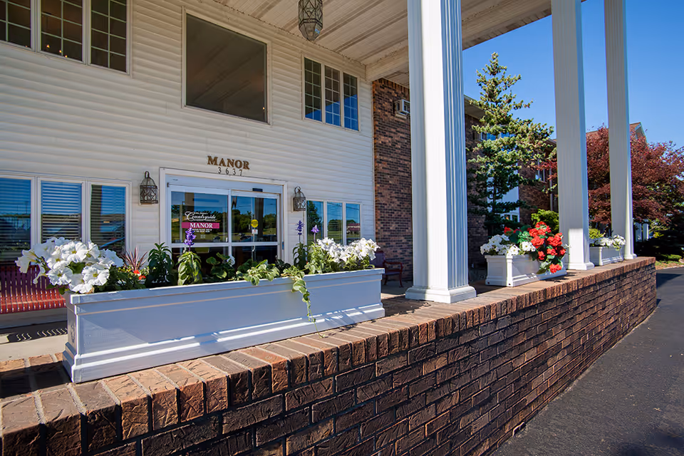 Entrance of Countryside Manor featuring a white building facade with large windows, a glass door with the facility's name, decorative lanterns on either side, white flower boxes with blooming flowers, and tall white columns supporting a covered porch.