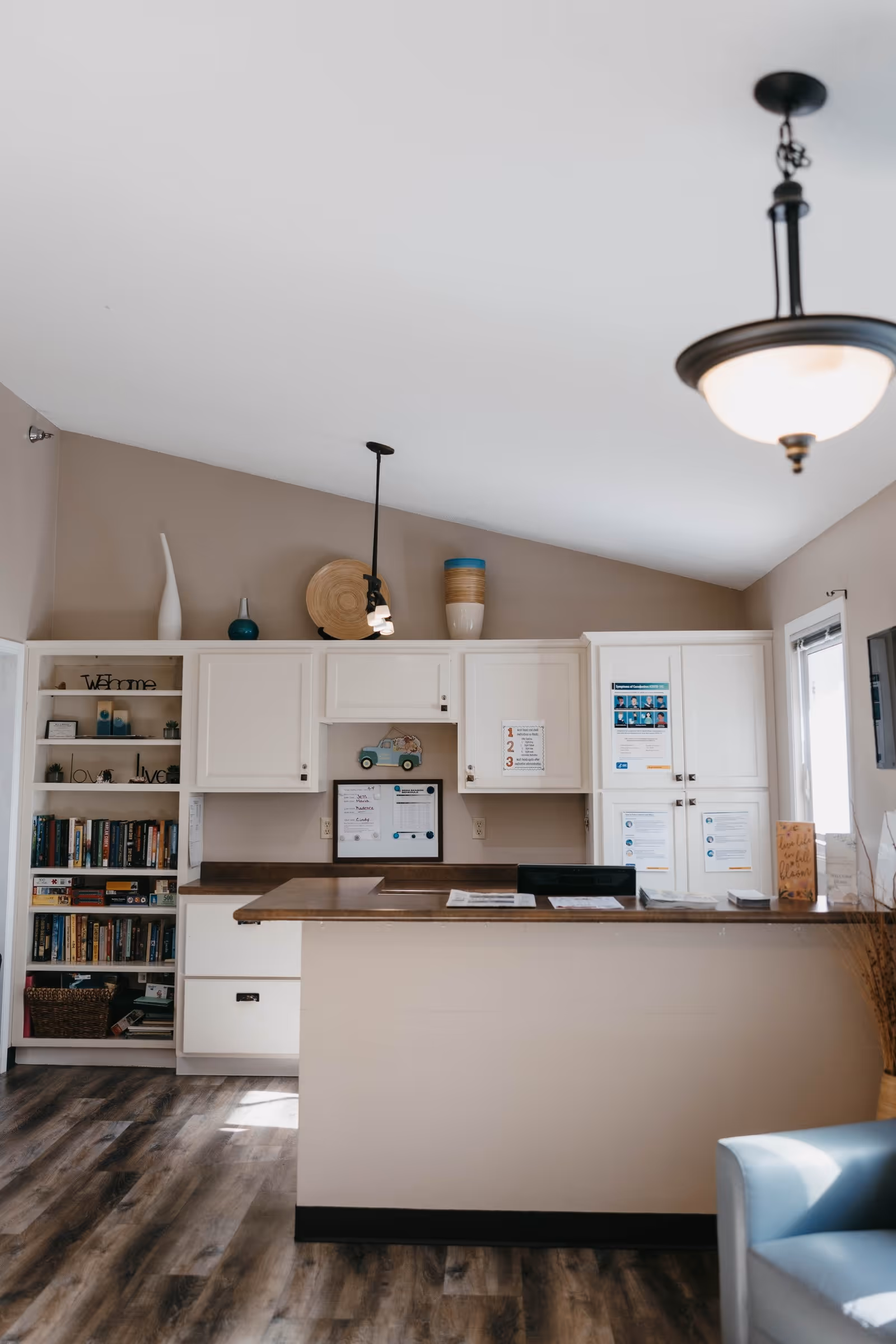 A reception or front desk area in a senior living facility with white cabinets, a wooden countertop, and shelves filled with books and decorative items. There are hanging pendant lights and a ceiling light fixture, with a light blue chair partially visible in the foreground.