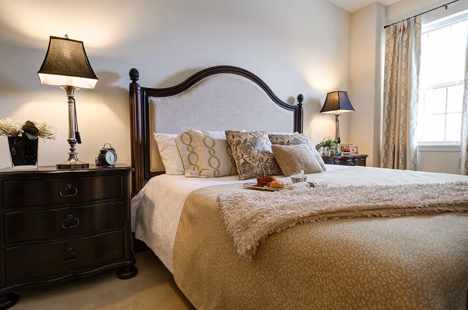 A cozy bedroom with a large bed featuring a beige upholstered headboard and multiple decorative pillows. There are two dark wooden nightstands on either side of the bed, each with a lamp and decorative items. A window with patterned curtains allows natural light to brighten the room.