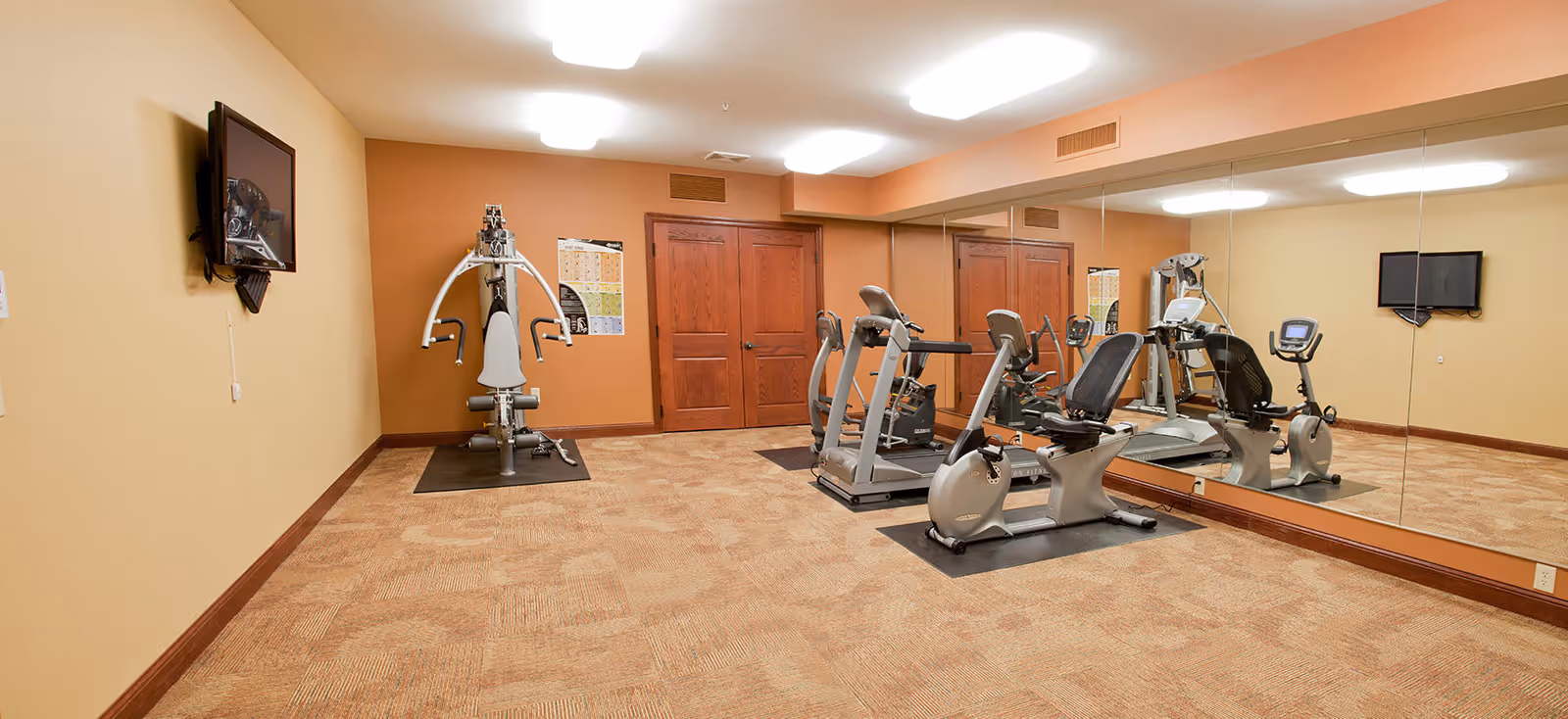 A senior living facility exercise room with beige walls and carpeted floor. The room contains various exercise equipment including a multi-gym machine, a recumbent exercise bike, and a treadmill. A large mirror covers one wall, reflecting the equipment. Two wooden doors are visible on the far wall, and a flat-screen TV is mounted on the wall opposite the mirror.