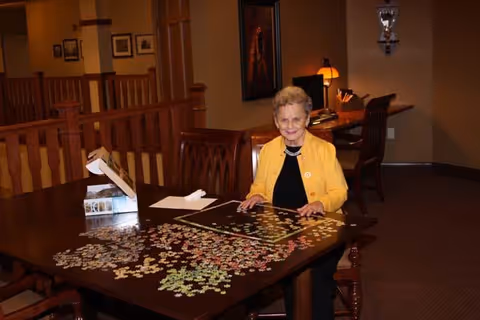 An elderly woman wearing a yellow cardigan is sitting at a wooden table working on a jigsaw puzzle in a warmly lit room with wooden furniture and framed pictures on the walls.