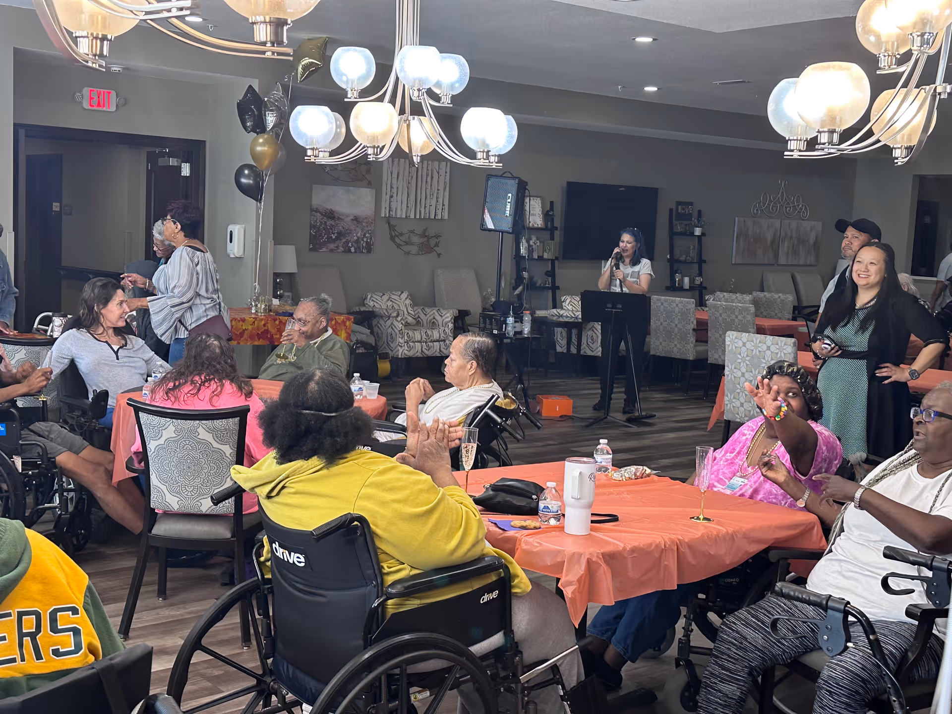Seniors seated at tables and in wheelchairs in a decorated communal dining/activity room watching a singer perform.
