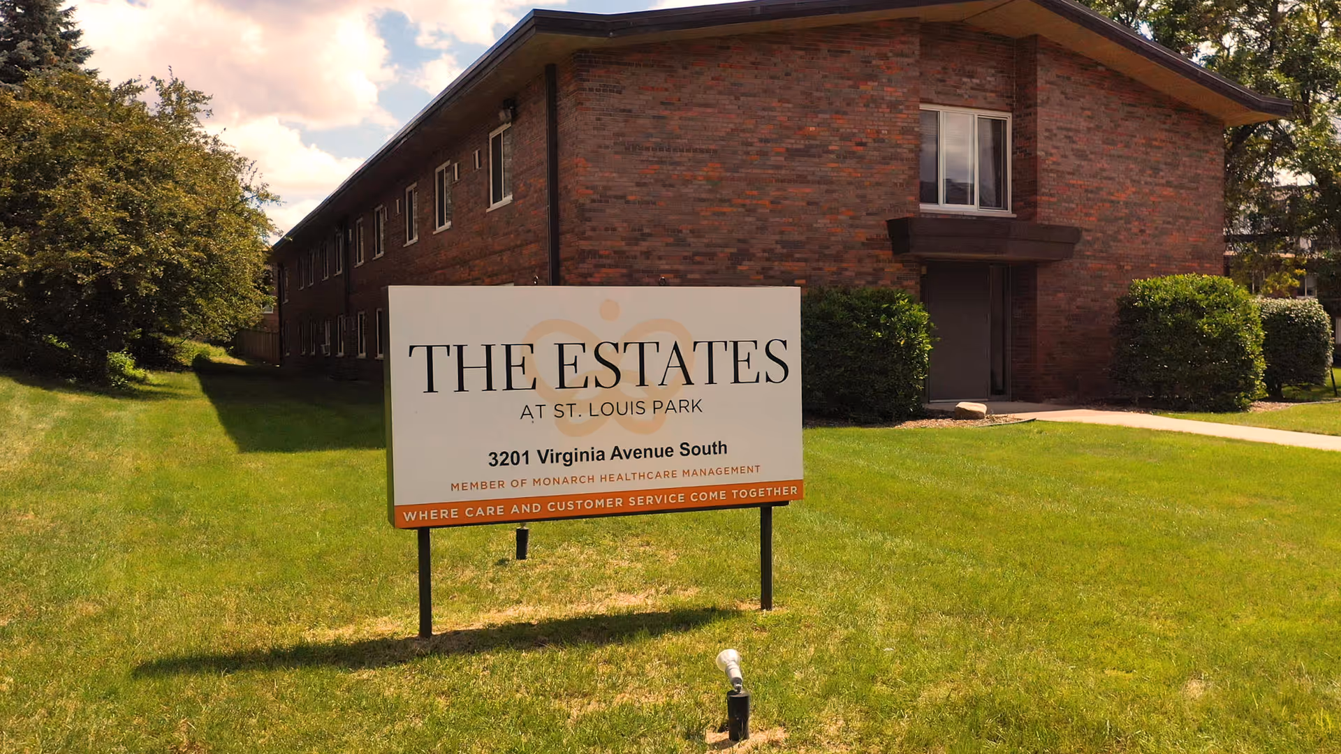 A brick building with multiple windows and a door, surrounded by green grass and bushes. In front of the building is a sign that reads 'THE ESTATES AT ST. LOUIS PARK, 3201 Virginia Avenue South, Member of Monarch Healthcare Management, Where Care and Customer Service Come Together.'