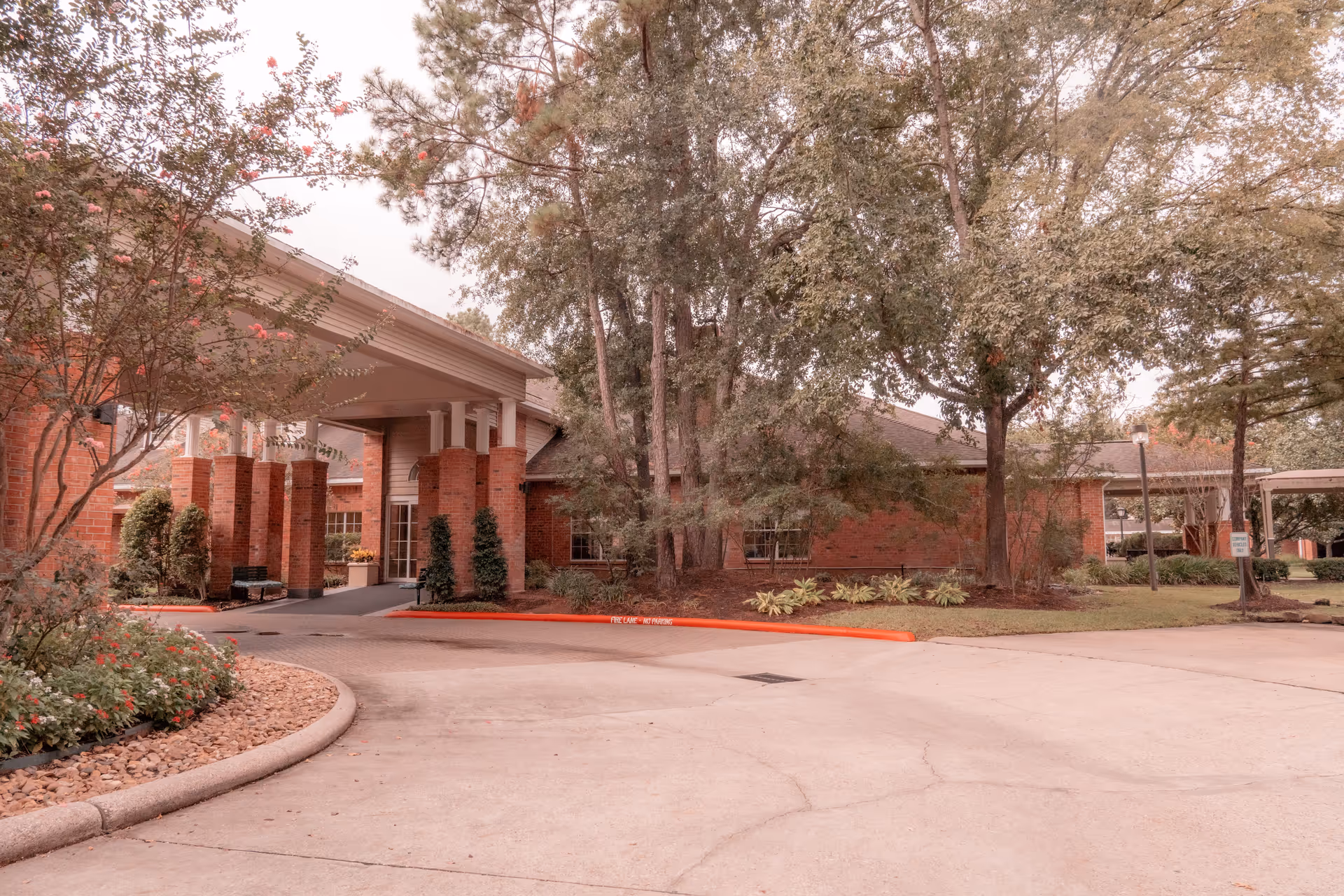Covered porte-cochère and entrance to a brick senior living building surrounded by trees and landscaping.