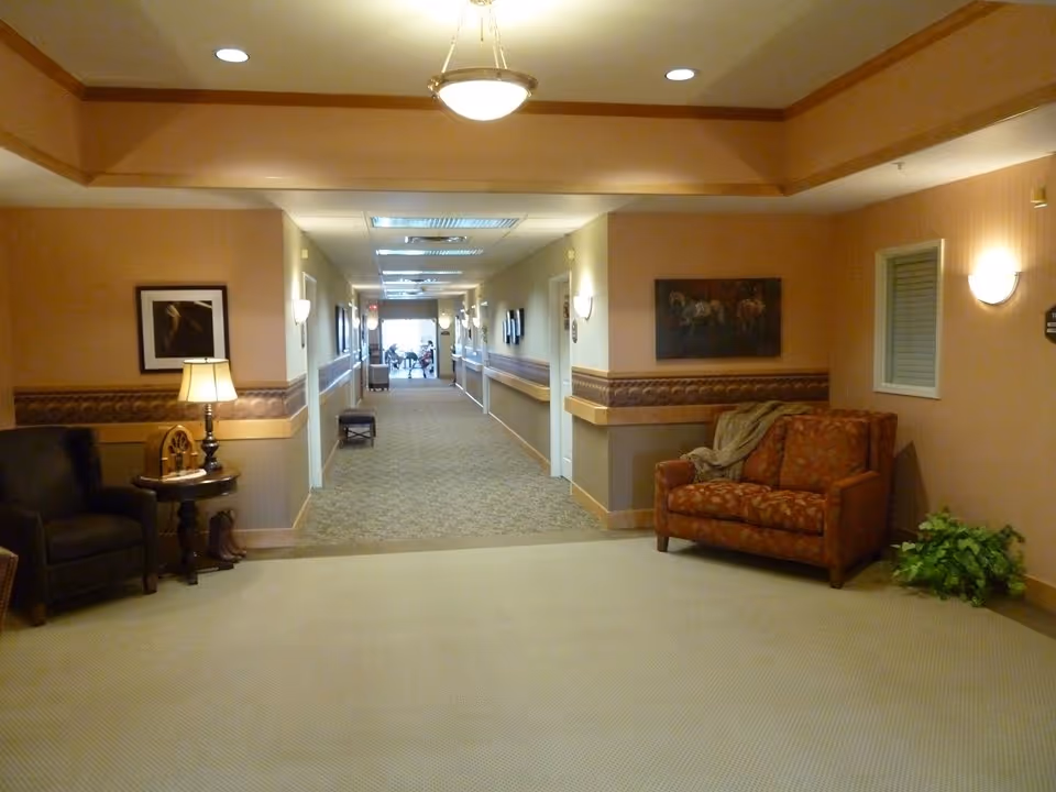 A warmly lit hallway in a senior living facility with beige walls and carpet. The hallway features a seating area with a patterned armchair, a small table with a lamp, framed artwork on the walls, and a plant in the corner. The corridor extends into the distance with additional seating and lighting fixtures visible.