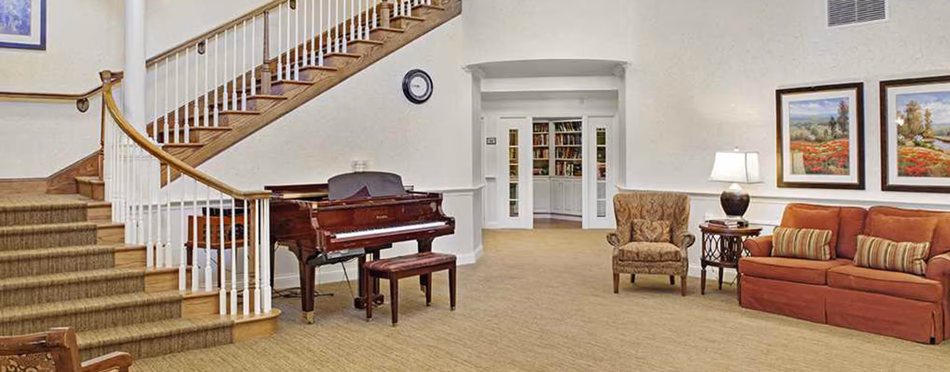 Spacious senior living common area with a grand piano by a curved staircase, seating with a sofa and armchair, and a doorway into a library.