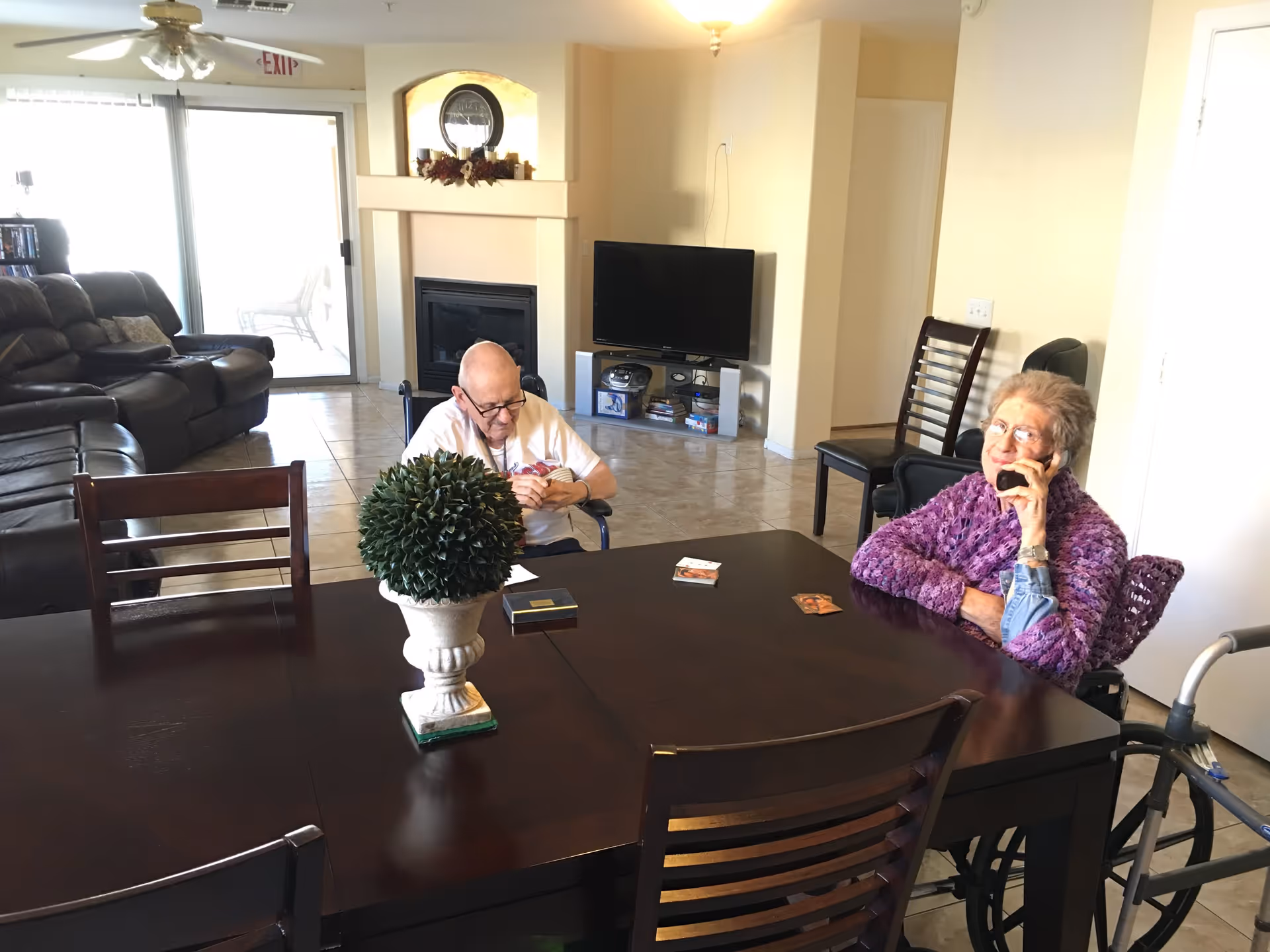 A living room and dining area in a senior living facility with two elderly people sitting at a dark wooden table. One person is in a wheelchair wearing a purple sweater and talking on the phone, while the other person is seated and looking down at something in their hands. The room has a tiled floor, a fireplace, a TV on a stand, and a sliding glass door leading outside. There is a decorative plant on the table.