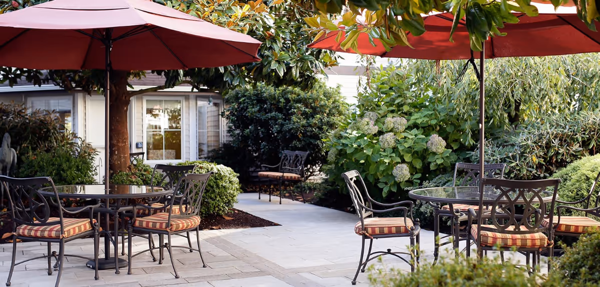 Outdoor patio area with round glass tables and metal chairs with striped cushions, shaded by large red umbrellas. The patio is surrounded by lush green bushes and plants, with a building and windows visible in the background.