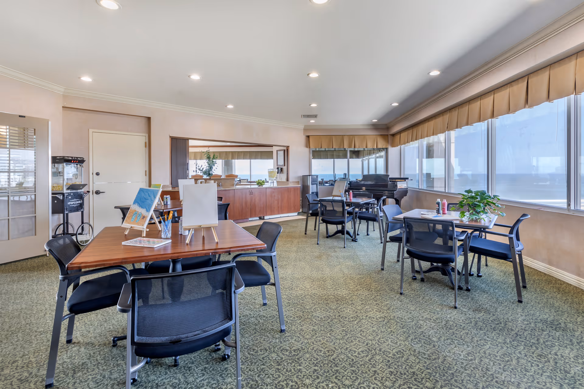 A bright activity room with several tables and chairs arranged for group activities. On one table, there are small easels with paintings and art supplies. Large windows line one side of the room, allowing natural light to fill the space. A piano is visible in the corner near the windows. The room has a patterned carpet and recessed ceiling lights.