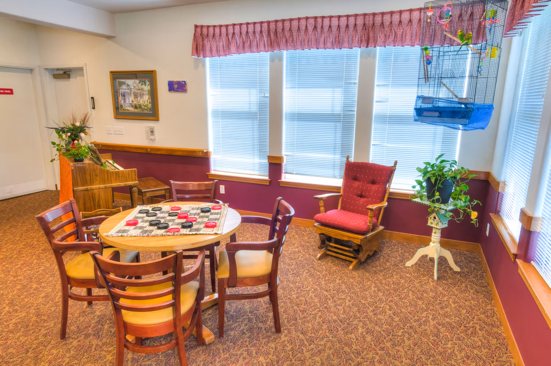 A cozy room with a round wooden table and six chairs arranged around it. On the table is a checkers board with red and black pieces. To the right, there is a red cushioned rocking chair and a small white plant stand holding a green potted plant. A birdcage with colorful toys hangs near the window. The room has large windows with blinds and a pink valance, carpeted floor, and walls painted white and maroon. A piano with a plant on top is visible in the background along with framed artwork on the wall.