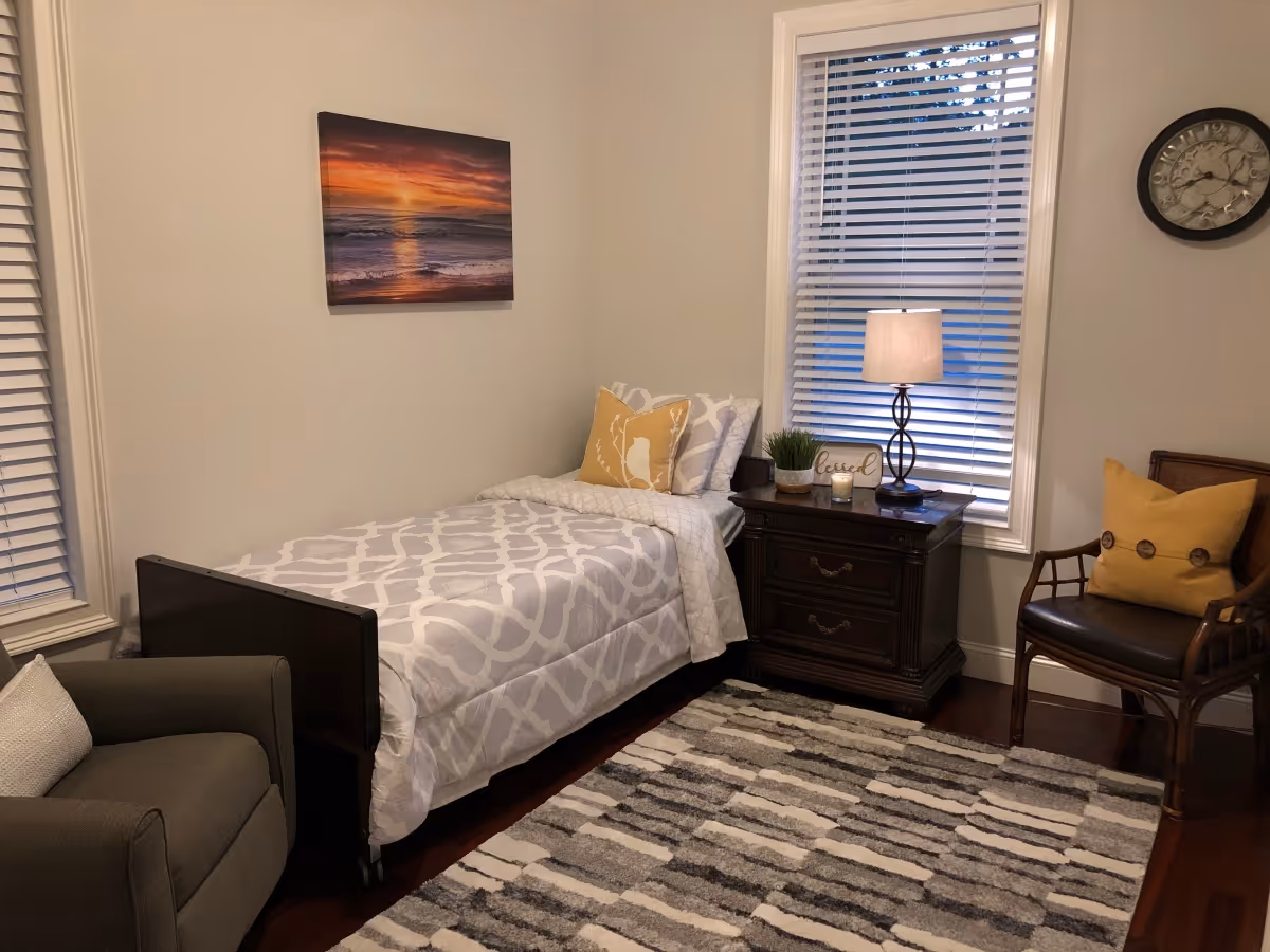 A cozy bedroom with a single bed covered in a white and gray patterned comforter, a yellow decorative pillow, and a nightstand with a lamp, small plant, and candle. There is a gray armchair with a white pillow, a wooden chair with a yellow pillow, a wall clock, and a framed sunset picture on the wall. The room has two windows with white blinds and a gray and white patterned rug on the wooden floor.
