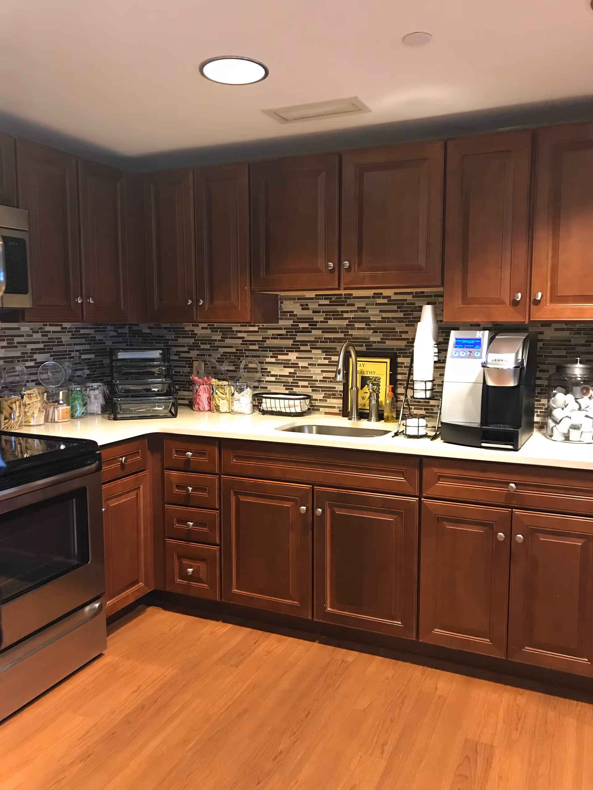 A kitchen with wooden cabinets, a stainless steel stove, a sink with a faucet, a coffee maker, and various containers with coffee supplies on the countertop. The backsplash features a mosaic tile design in neutral colors, and the floor is wood.